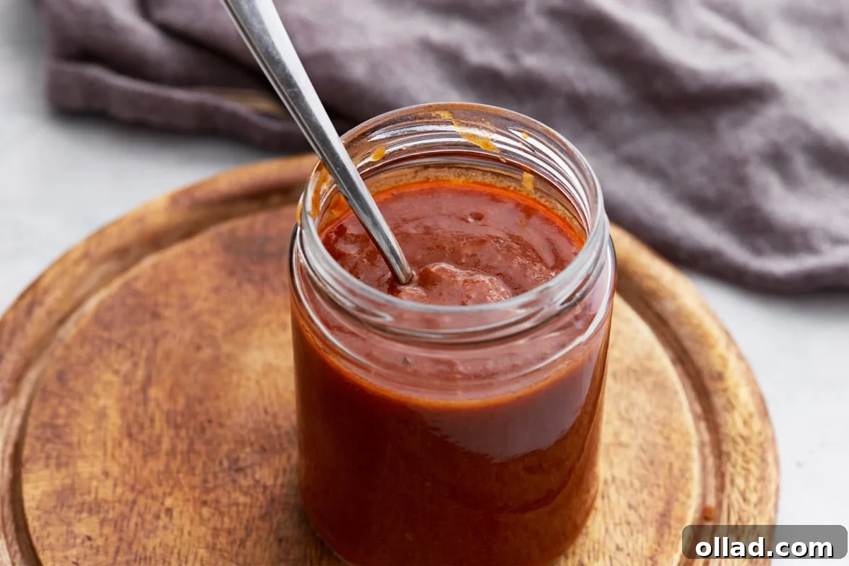 Side view of a glass jar of enchilada sauce on a round wooden board with a spoon inside.