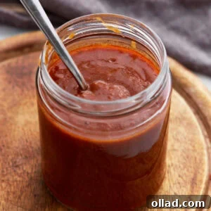 A glass jar filled with rich, dark red enchilada sauce with a spoon inside, sitting on a wooden board.