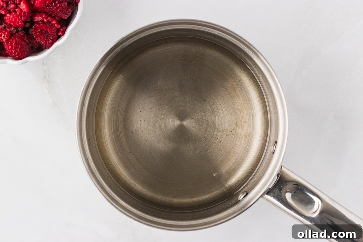 A silver saucepan with clear simple syrup gently simmering on a stovetop, ready for the next step of making raspberry sorbet.
