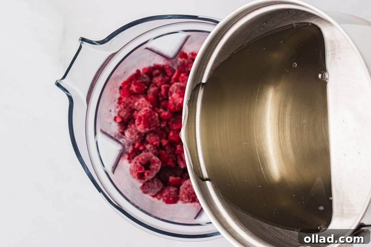 A saucepan of freshly made simple syrup being poured into a blender filled with vibrant frozen raspberries and lemon slices, preparing for blending.