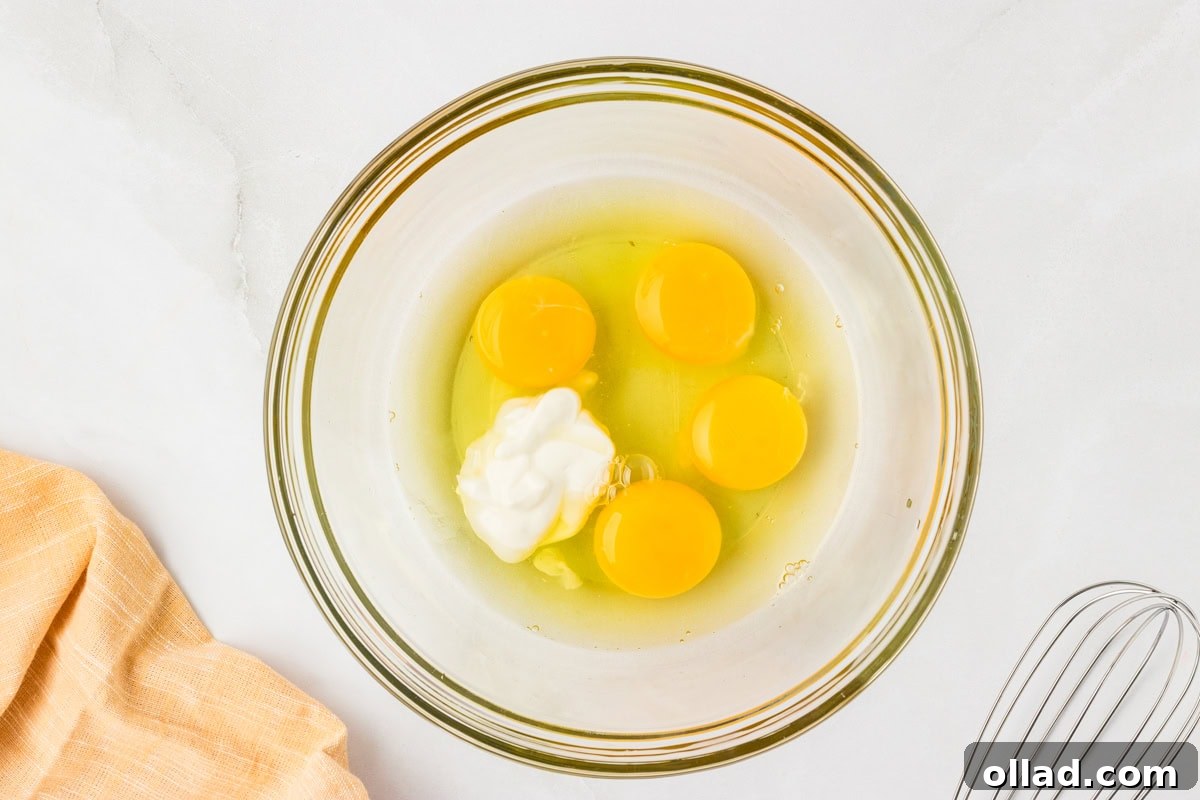 Ultimate Keto Cornbread 5 Mixing bowl with four cracked eggs and sour cream before whisking, sitting on a white counter with a whisk to the side.