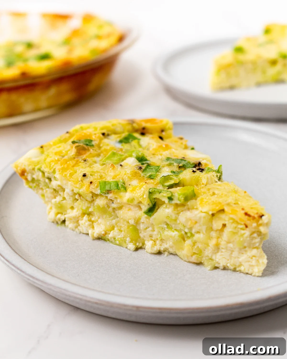 A vibrant slice of crustless zucchini quiche on a modern gray plate, garnished with fresh green onions. The full golden-brown quiche is softly visible in the background, promising culinary delight.