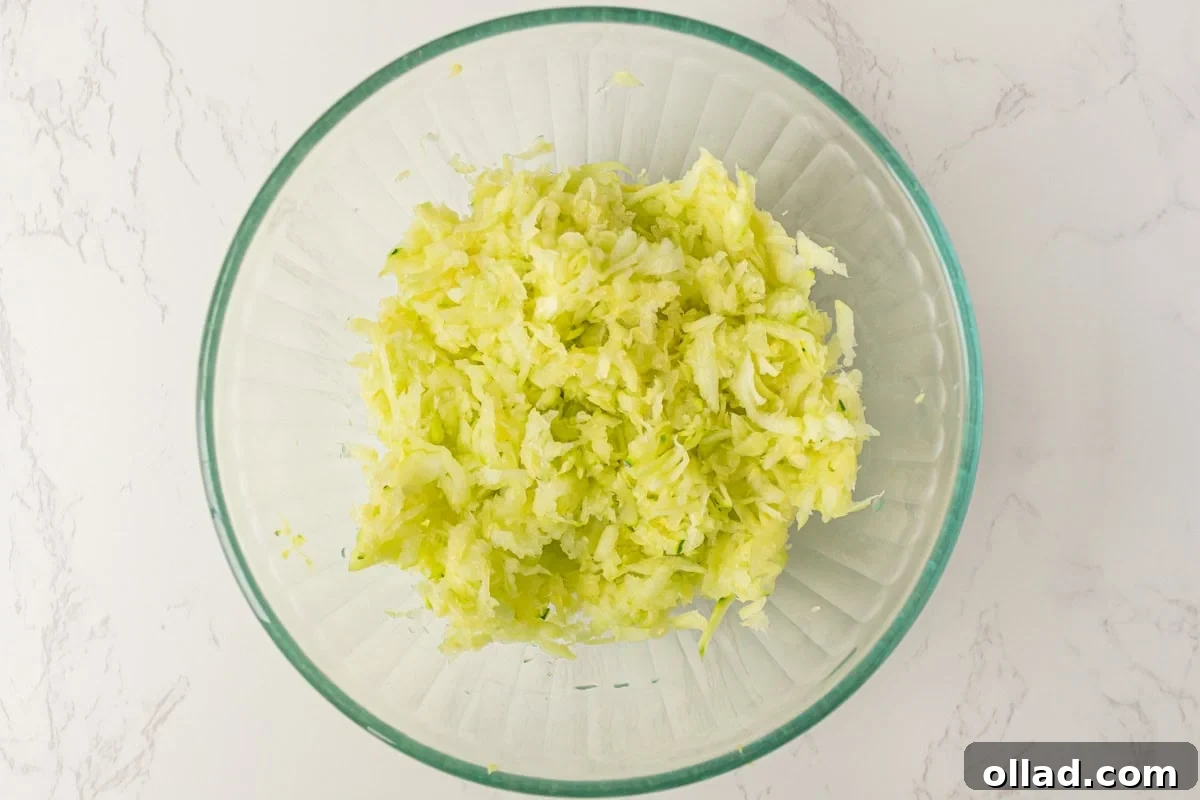 Freshly shredded zucchini resting in a clear glass mixing bowl on a sleek marble countertop, ready for the crucial step of moisture removal, highlighting its texture.