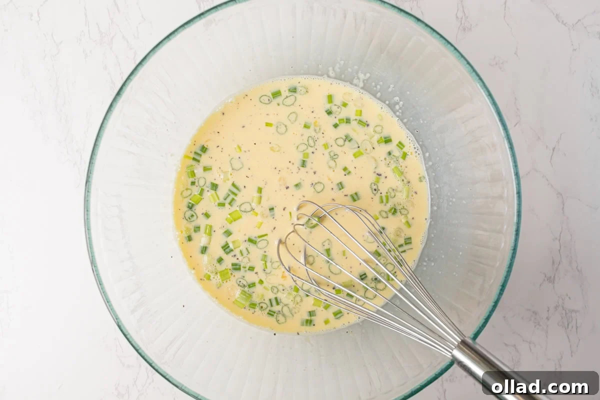 The smooth egg mixture, now combined with finely chopped green onions, a generous pour of half and half, salt, and pepper, all perfectly blended in a clear glass bowl, ready for the next step.