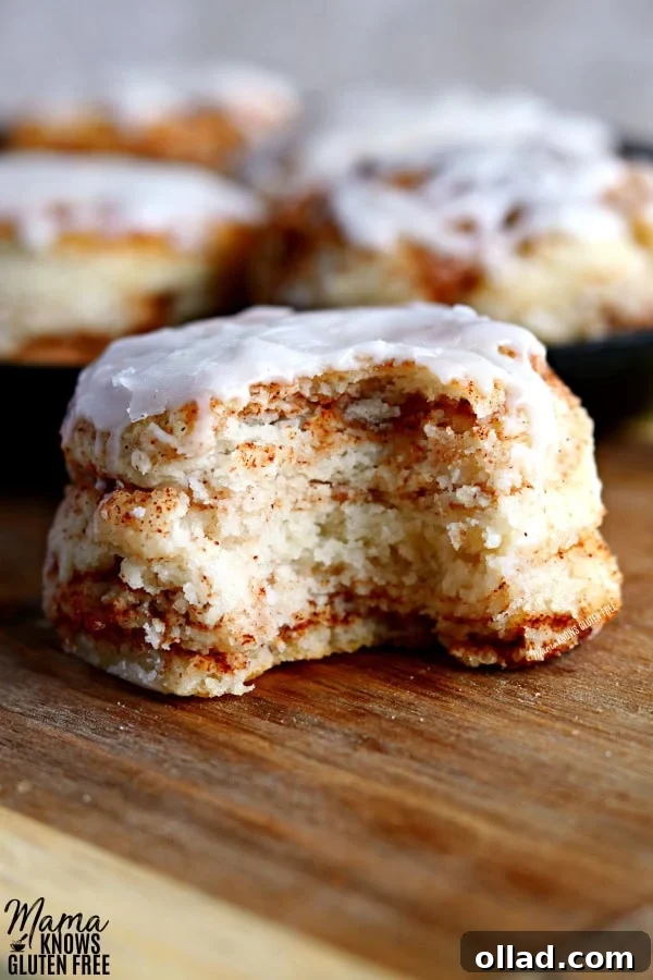 Warm Spiced Gluten-Free Biscuits, Dairy-Free Friendly 2 Tender and flaky gluten-free cinnamon biscuit with a bite taken out, showing the layered texture and rich cinnamon swirl. More golden biscuits are visible in a rustic cast iron skillet in the background, topped with a glistening vanilla glaze.