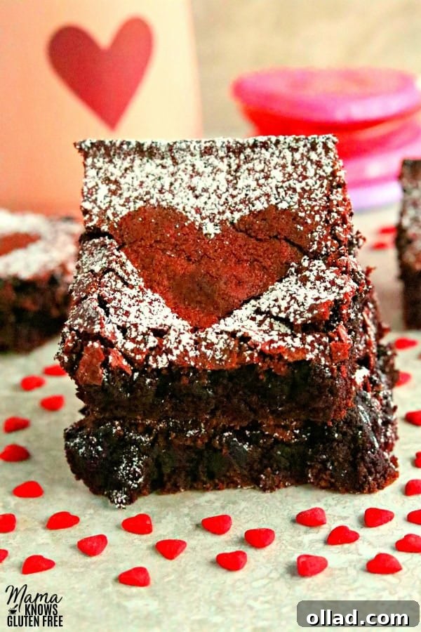 Fudgy Gluten-Free Dairy-Free Red Velvet Brownies 2 Two gluten-free red velvet brownies stacked with more brownies, a heart cup, and heart candy in the background, signifying a sweet indulgence.