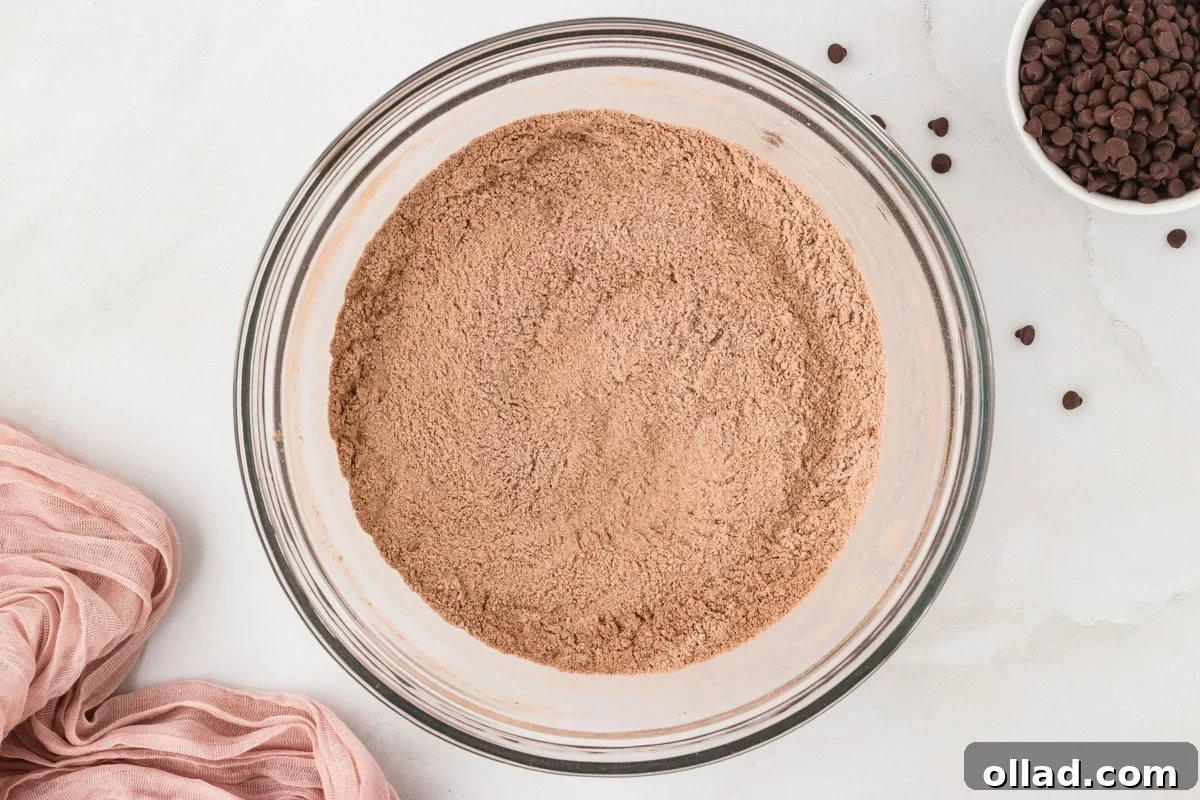 Decadent Gluten-Free Double Chocolate Muffins 5 Glass bowl filled with thoroughly blended dry ingredients for chocolate muffin batter, showing cocoa powder and flour mixed evenly, with chocolate chips and a pink cloth nearby.