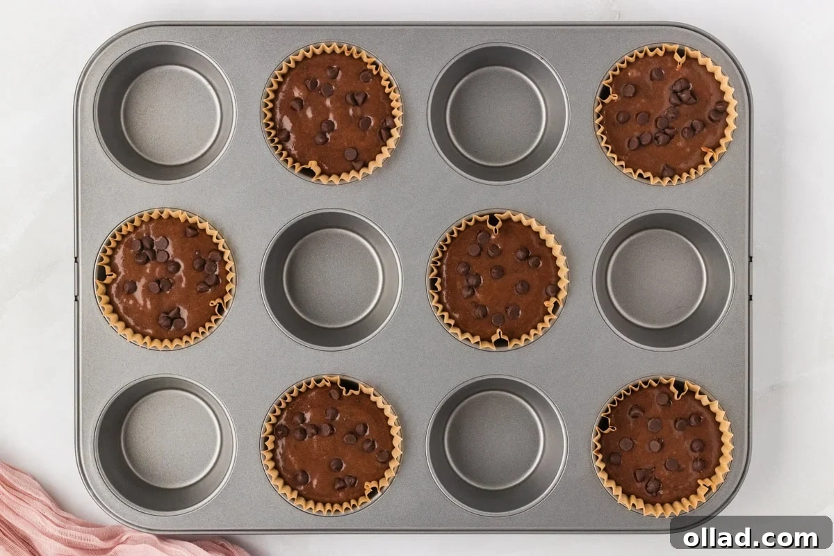 Decadent Gluten-Free Double Chocolate Muffins 8 Chocolate muffin batter portioned neatly into paper-lined muffin cups in a metal tin, topped with additional chocolate chips, ready for oven baking.