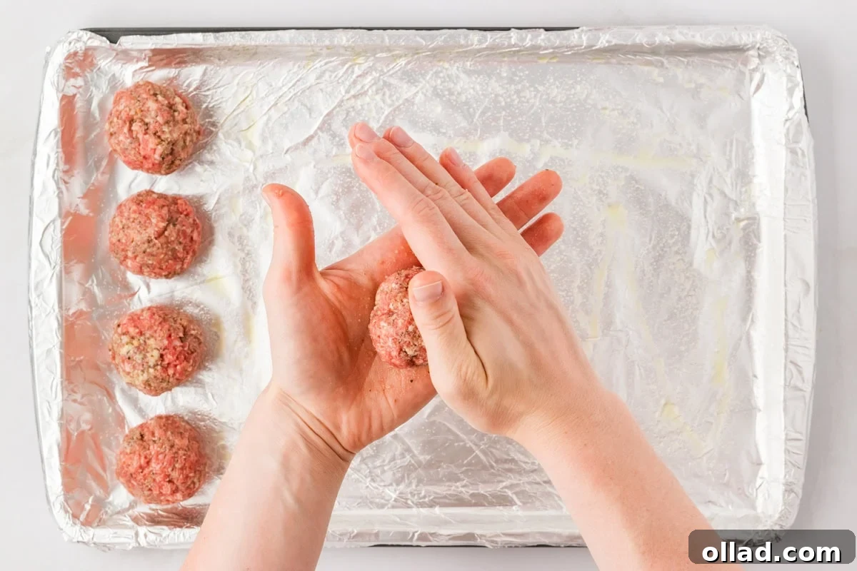 Delicious Gluten-Free Meatballs 9 Hands shaping a meatball over a foil-lined baking sheet with already formed meatballs in the background.