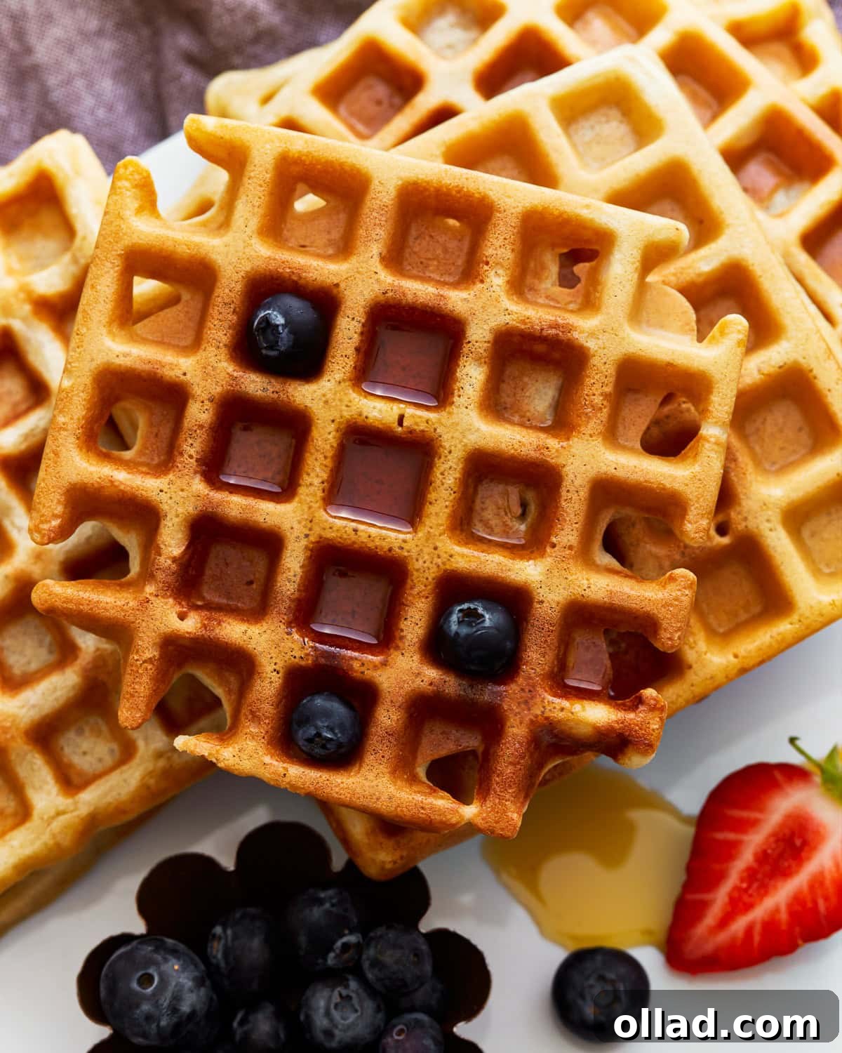 A beautifully stacked plate of golden brown gluten-free waffles, generously drizzled with maple syrup and sprinkled with fresh blueberries. A perfect, inviting breakfast scene.