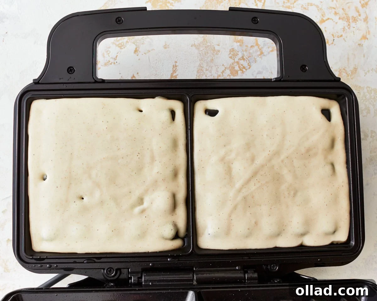 Gluten-free waffle batter being carefully poured from an ice cream scoop into a hot, greased waffle iron.