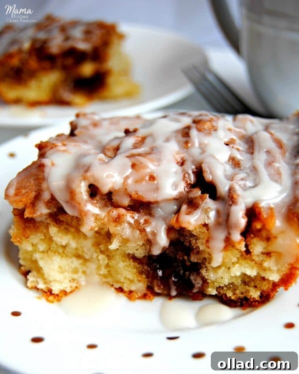 slice of gluten-free cinnamon roll cake on a white plate with another slice and coffee cup in the background