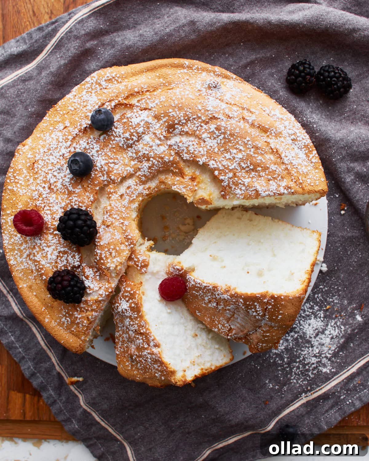 Cloud-Light Gluten-Free Angel Food Cake 2 A perfectly sliced gluten-free angel food cake resting on a grey towel, garnished with powdered sugar and fresh berries, ready to be enjoyed.