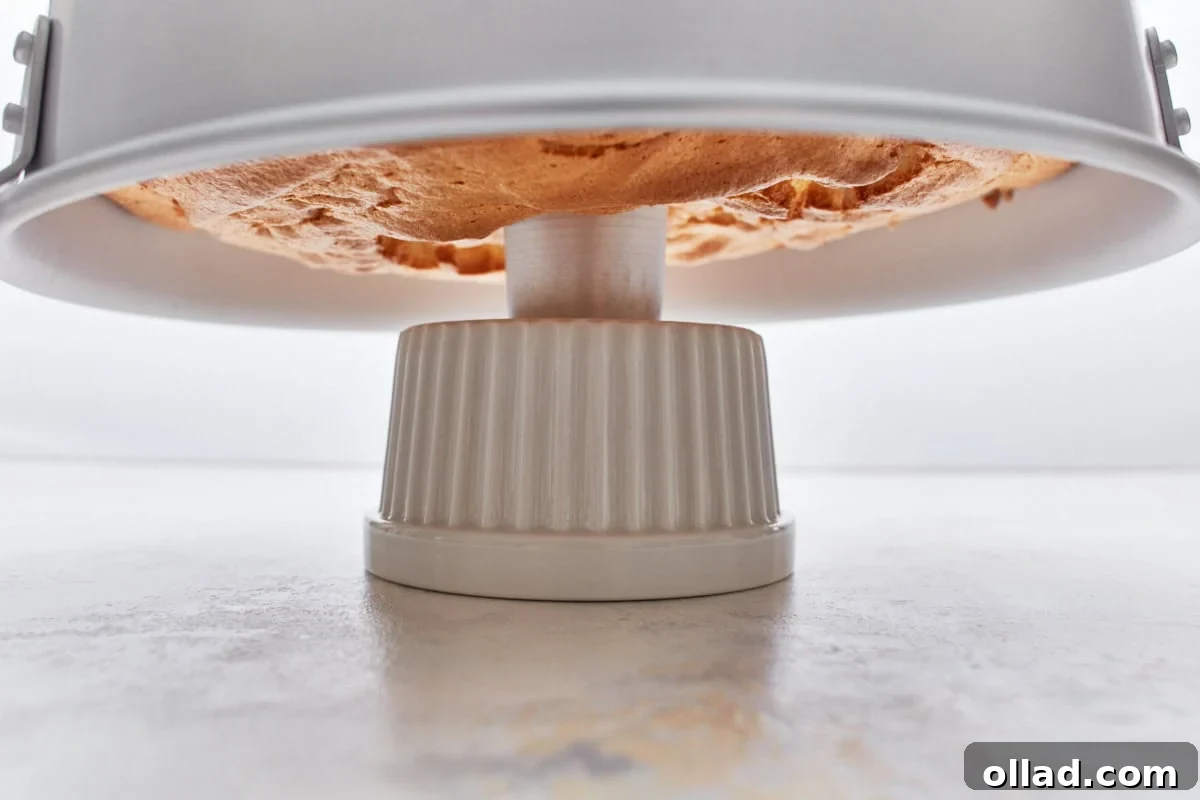 Cloud-Light Gluten-Free Angel Food Cake 11 A freshly baked gluten-free angel food cake in its tube pan, inverted and cooling upside down on a ceramic holder to prevent collapse.