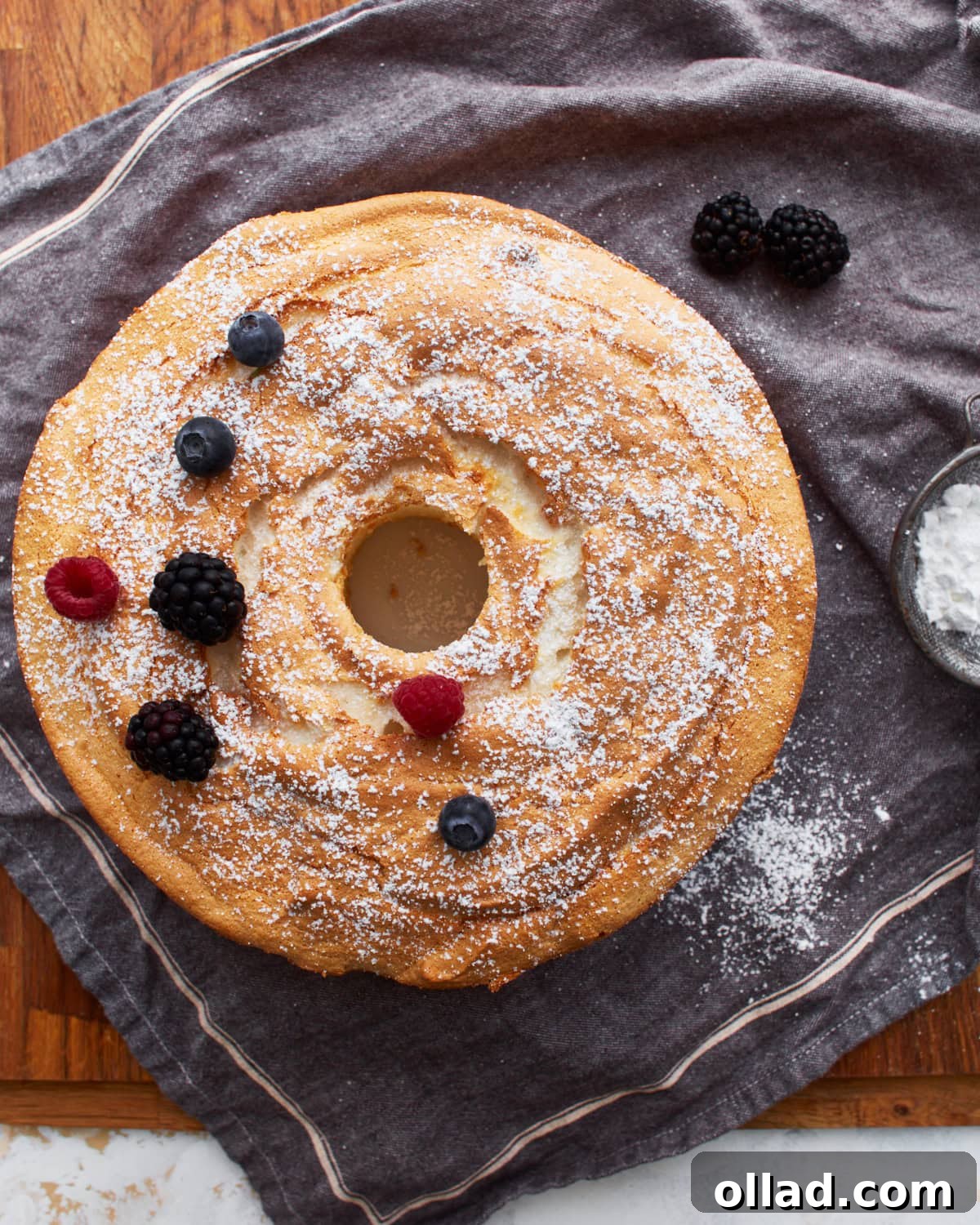 Cloud-Light Gluten-Free Angel Food Cake 16 A pristine, whole gluten-free angel food cake resting on a grey towel, elegantly dusted with powdered sugar and adorned with a scattering of fresh blackberries, raspberries, and blueberries.
