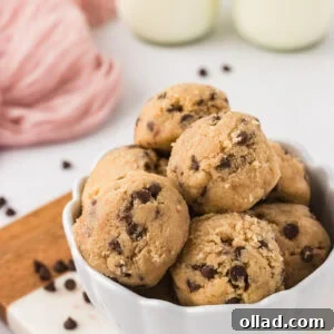 A rustic wooden bowl brimming with scoops of golden-brown gluten-free edible chocolate chip cookie dough, garnished with scattered mini chocolate chips. Two tall glasses of cold milk are artfully blurred in the background, completing a comforting scene of sweet indulgence.