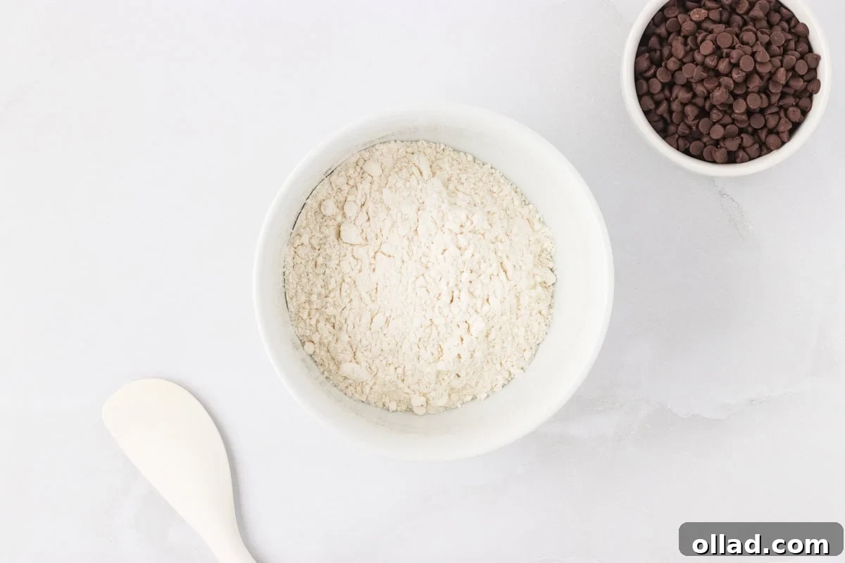 Irresistible Gluten-Free Cookie Dough 5 A white bowl filled with gluten-free flour sits prominently next to a smaller bowl brimming with mini chocolate chips, all arranged on a pristine white surface. A spatula rests nearby, awaiting its role in mixing.