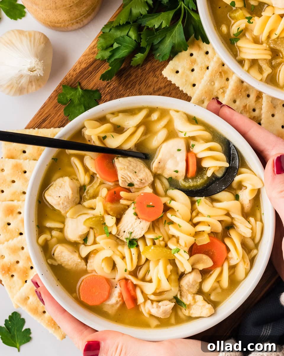 A pair of hands gently cradling a warm, inviting bowl of gluten-free chicken noodle soup. The soup is generously filled with tender chicken pieces, spiral noodles, carrots, and celery, beautifully topped with fresh parsley. In the background, crispy crackers and additional fresh herbs are visible, creating a comforting and appetizing scene.