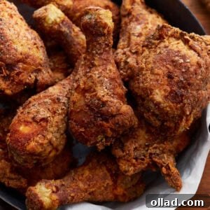 A stack of perfectly crispy, golden-brown gluten-free fried chicken pieces rests invitingly in a black skillet, which is lined with a paper towel and placed on a rustic wooden surface. The chicken boasts a rich, textured crust, promising a delightful crunch.