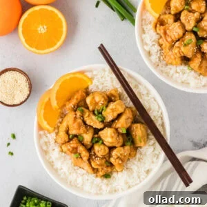 A white bowl filled with glistening gluten-free orange chicken, perfectly arranged over a bed of fluffy white rice. The dish is beautifully garnished with vibrant fresh orange slices, chopped green onions, and a sprinkle of sesame seeds, with whole oranges and scallions artfully placed in the soft-focus background.