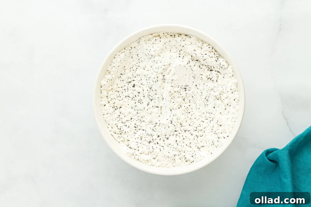 Tangy & Tender Gluten-Free Lemon Poppy Seed Muffins 5 Dry ingredients for gluten-free lemon poppy seed muffins, including gluten-free flour and poppy seeds, whisked together in a white bowl on a marble surface.