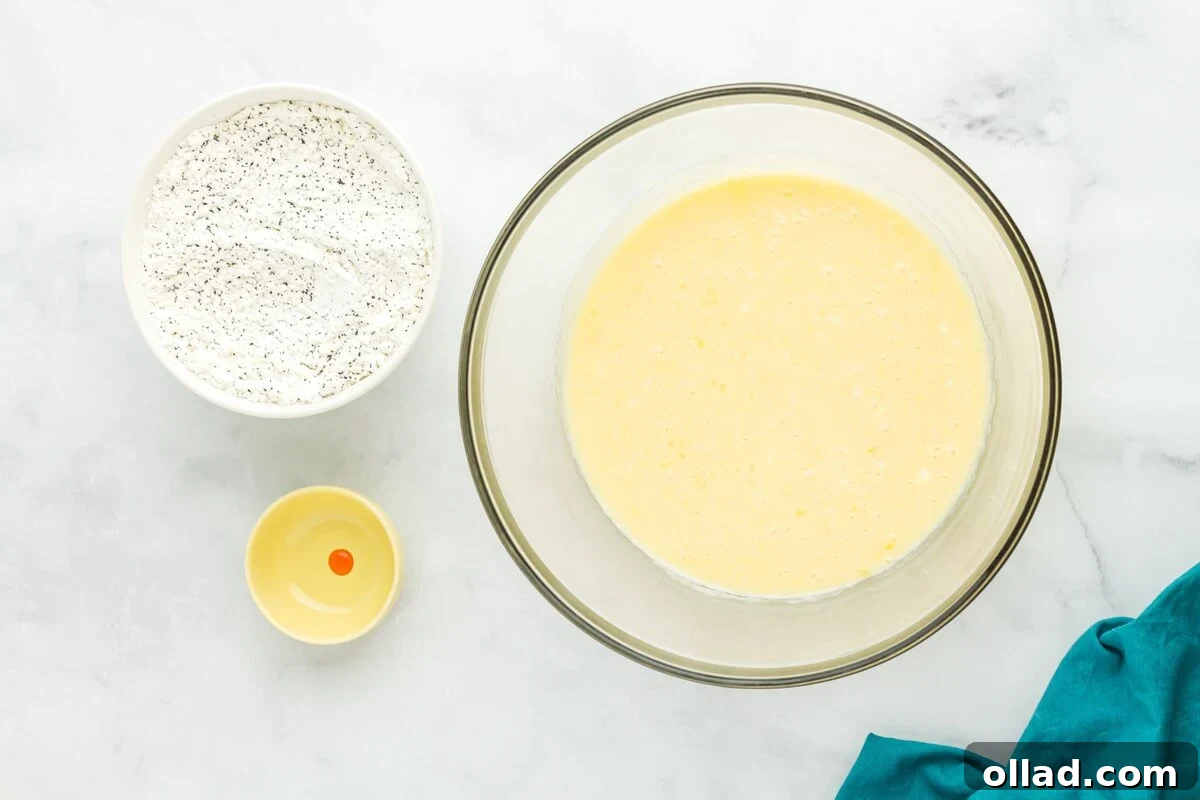 Tangy & Tender Gluten-Free Lemon Poppy Seed Muffins 6 Wet ingredients for gluten-free lemon poppy seed muffins, including sour cream, melted butter, eggs, lemon juice, and zest, mixed in a large glass bowl, with a small bowl of yellow food coloring nearby.