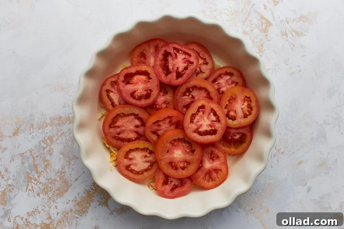 Layer of sliced tomatoes arranged over the cheese mixture in a white pie dish before baking.