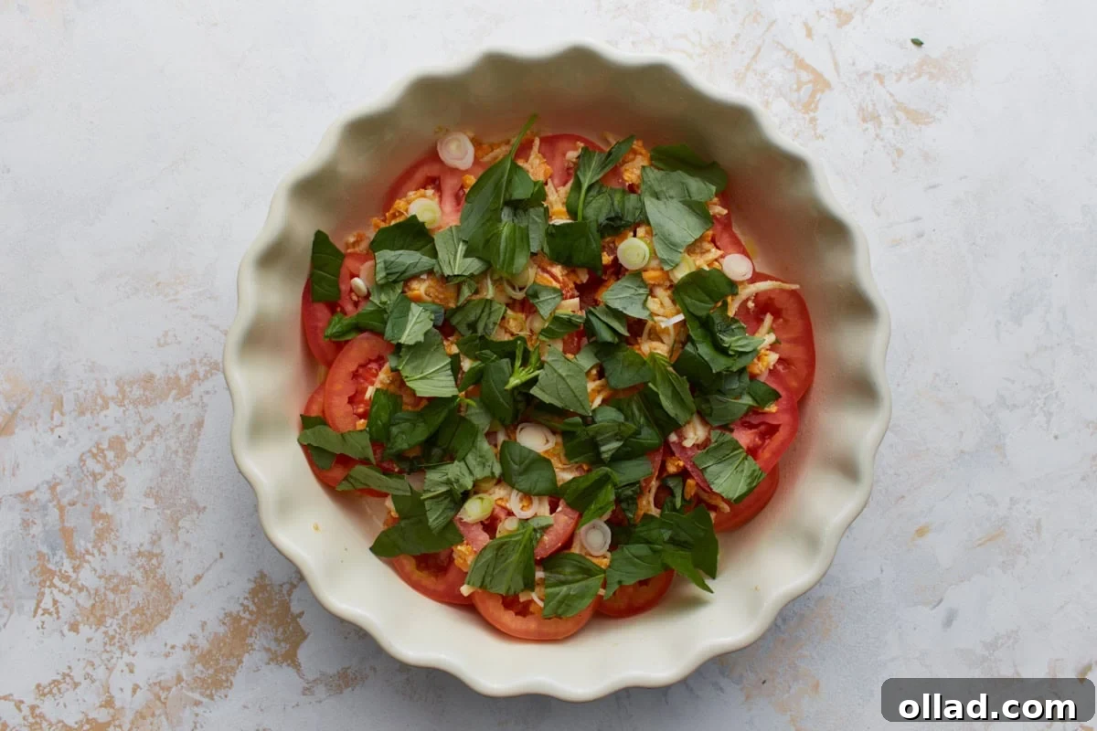 Overhead view of crustless tomato pie before baking with layers of sliced tomatoes, cheese mixture, fresh basil, and green onions in a white pie dish.