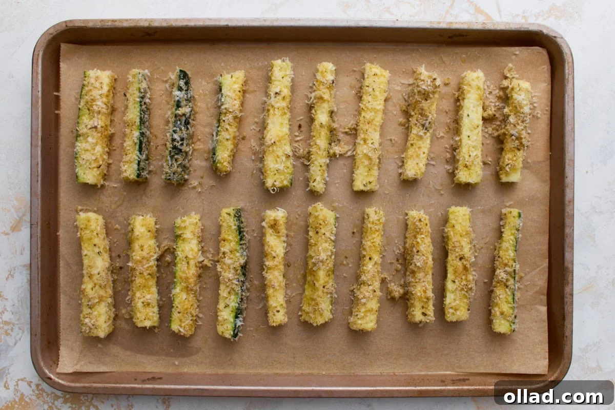 Crispy Keto Zucchini Fries: Low-Carb Snack Recipe 10 Breaded zucchini fries arranged in a single layer on a parchment-lined baking sheet before baking.
