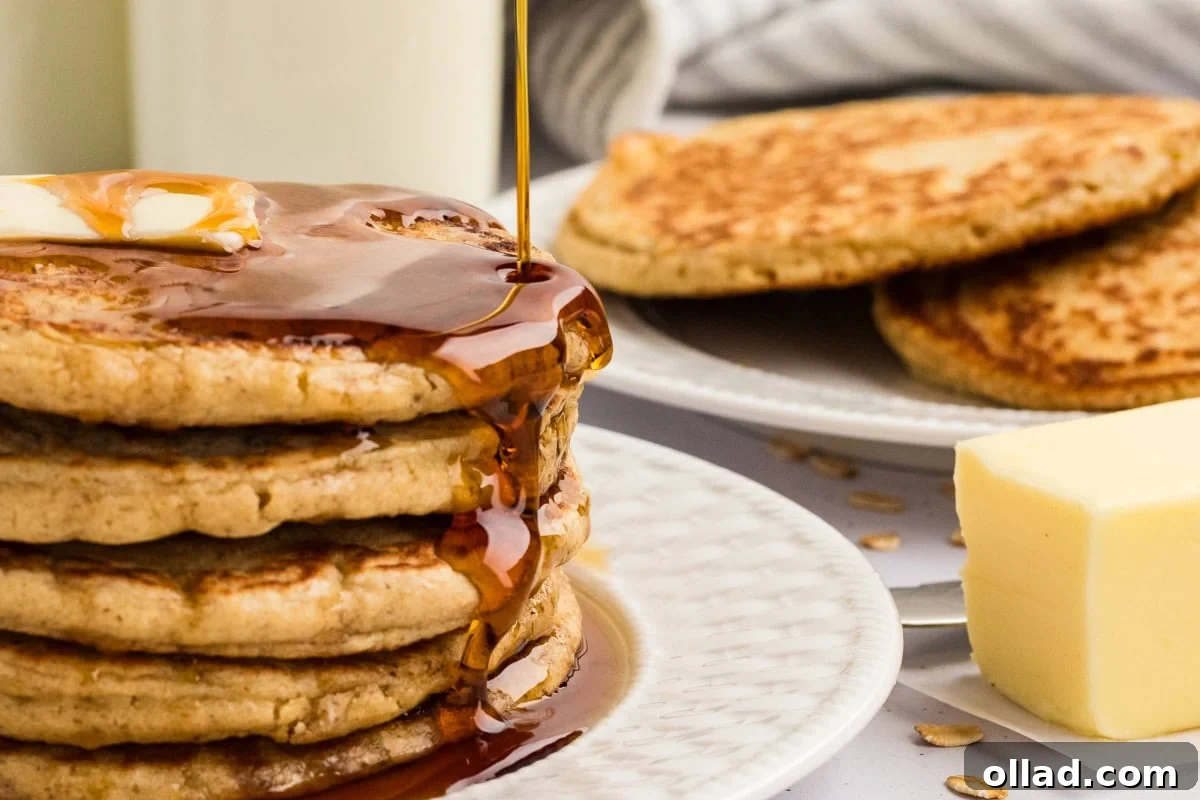 Maple syrup pouring over a stack of buttered oat flour pancakes.