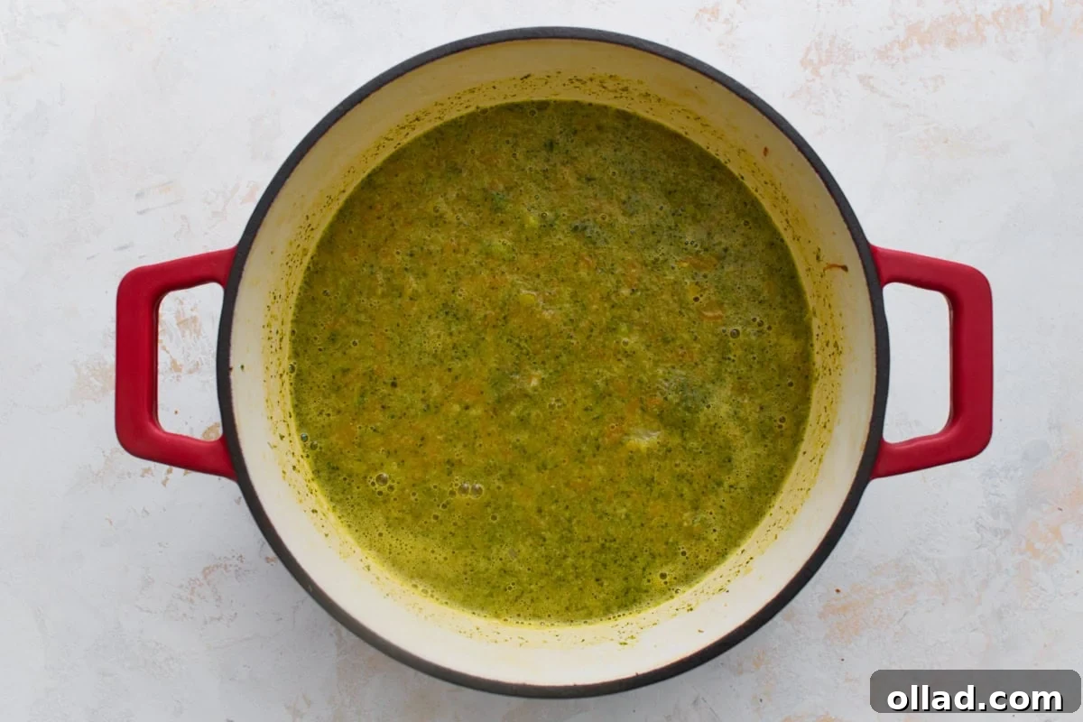 Broccoli, carrots, and broth simmered and partially blended into a chunky, bright green soup base in a red Dutch oven.