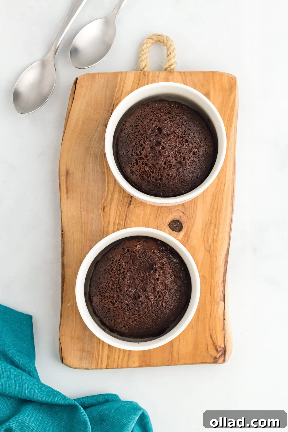 Two flourless mug cakes baked in white ramekins on a wooden board with spoons and a teal napkin