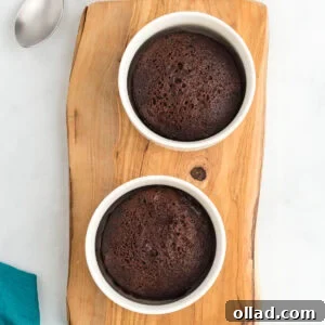 Two flourless mug cakes baked in white ramekins on a wooden board with spoons and a teal napkin